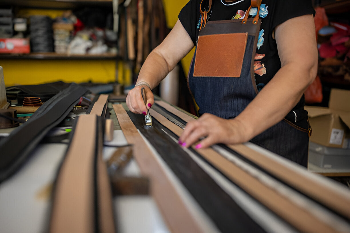 Artisan wearing a denim apron carefully applying finish to long leather strips on a workbench, surrounded by tools and materials in a workshop, highlighting precision and handcrafted leatherwork.